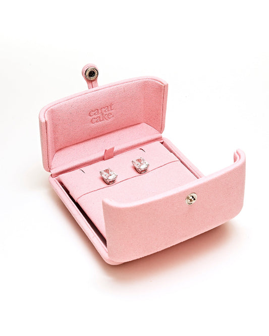 Pink jewelry box with square pair of diamond earrings inside on a white background